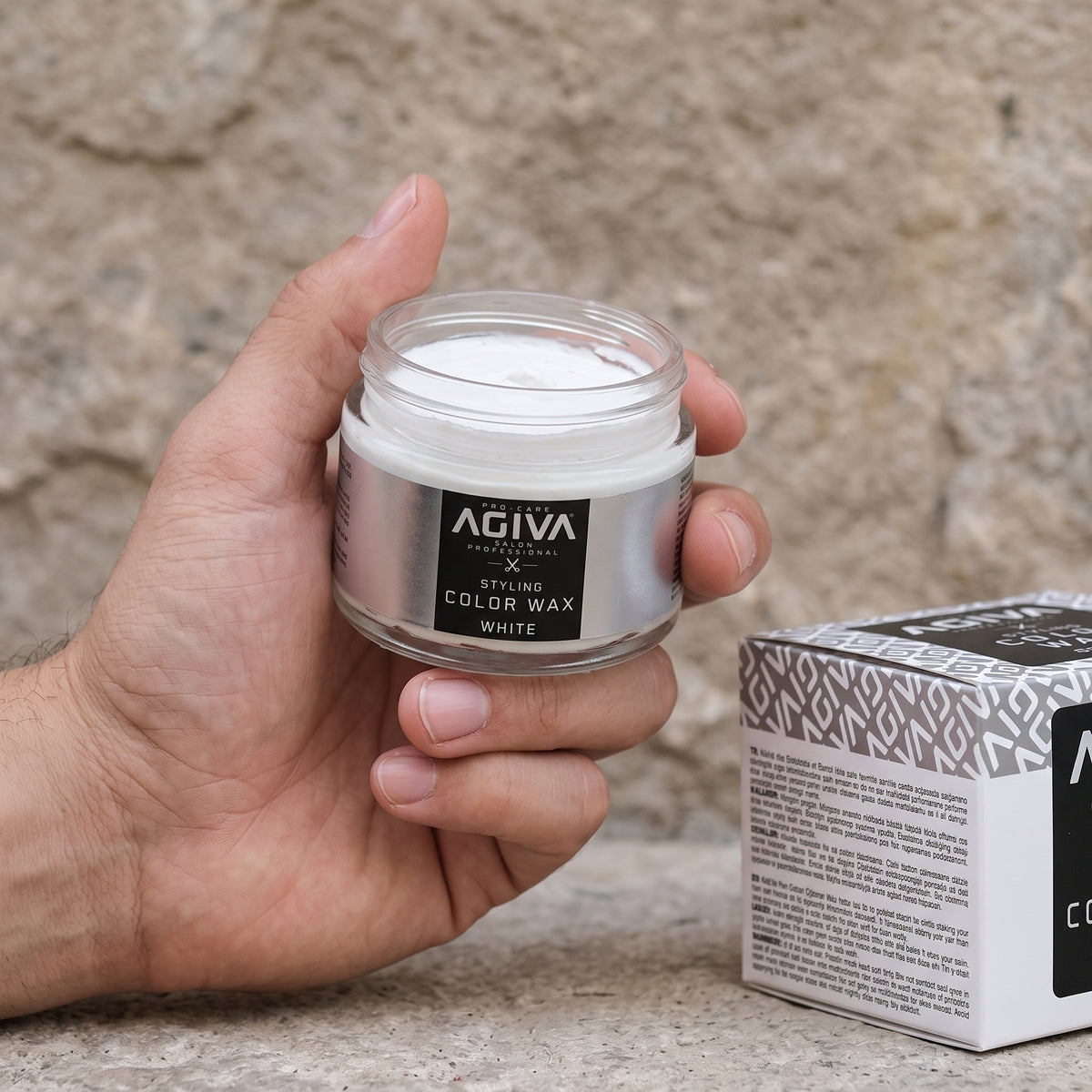 Hånd holder åben glaskrukke med Agiva Styling Color Wax White ved siden af æske på stenunderlag.
