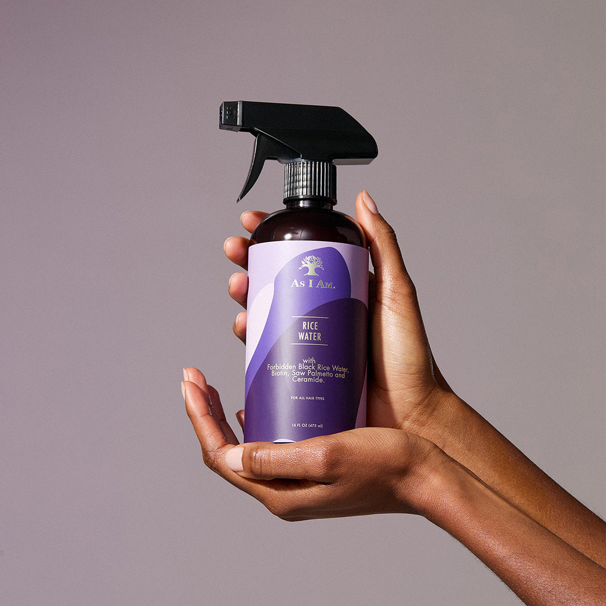 Hænder holder en flaske As I Am Rice Water Spray (475 ml) foran en lilla baggrund.