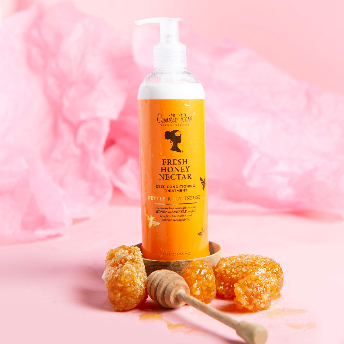 Hvid flaske med pumpe af Camille Rose Fresh Honey Nectar Deep Conditioning Treatment på en lyserød baggrund. Ved siden af flasken ligger honningkamme og en honningdipper.