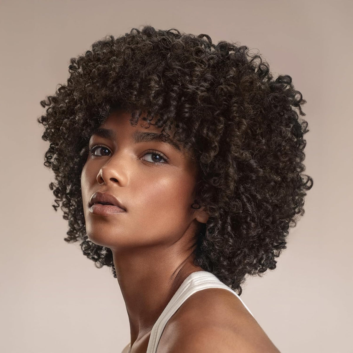 Kvinde med naturligt krøllet afro-frisure og curly bangs, iført hvid top mod beige baggrund.