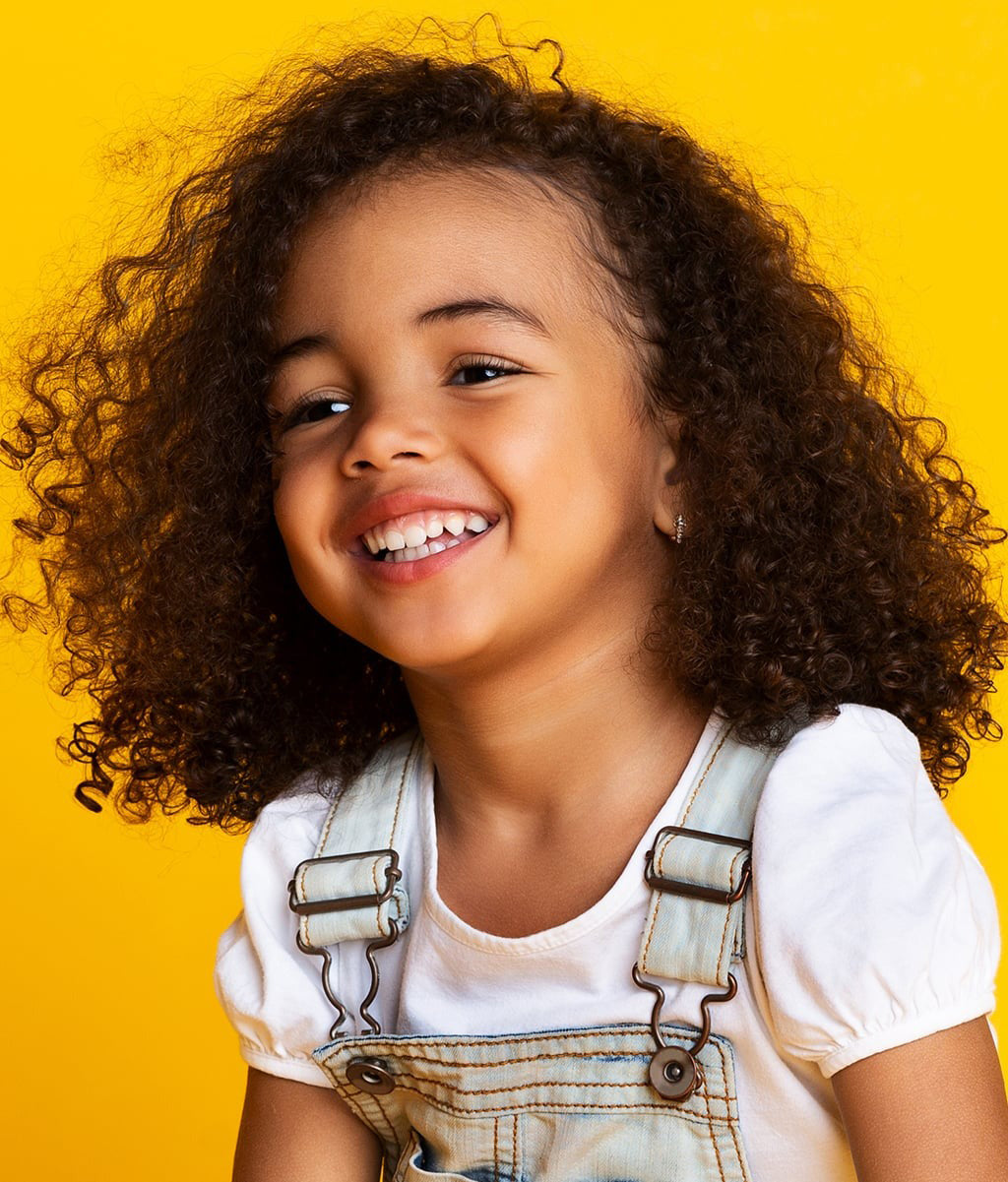 Smilende barn med krøllet hår, der fremstår sundt og velplejet med Not Your Mother's Kids Curly Care Shampoo 237ml.