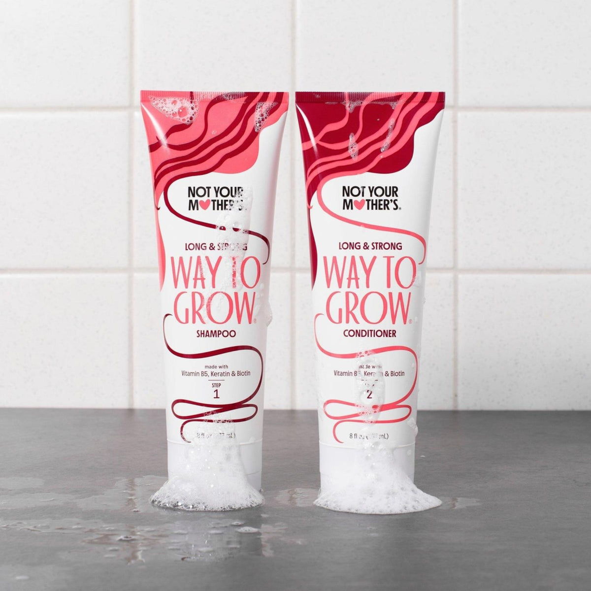 Not Your Mother's Way To Grow shampoo og conditioner i pink-hvide tuber med skum på grå overflade.